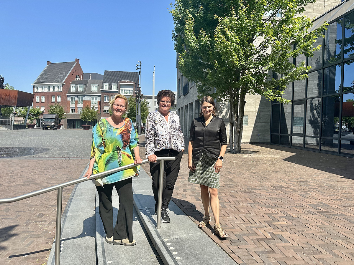 Wethouder Zeeman samen met wijk-GGD'er Petra Cooijmans en ondersteuner Patricia Smets op de trap van het bordes va het gemeentehuis met uitkijk op het zonnige Dorpsplein.