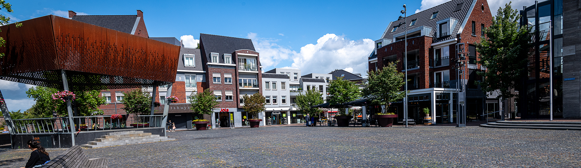 Wijds zicht op het Dorpsplein met de kiosk en de winkels op de Hoofdstraat met op de achtergrond een blauwe lucht met stapelwolken.